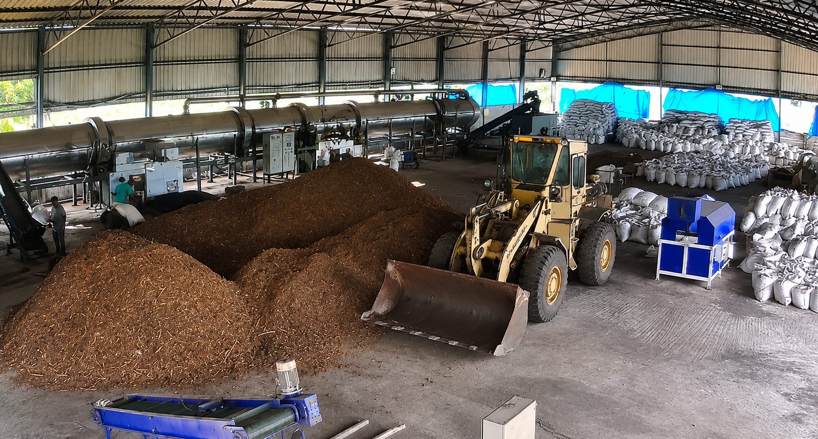 Wide view of the Kakinada line — rotary pyrolysis drum, feedstock handling by loader, and bagged biochar output.